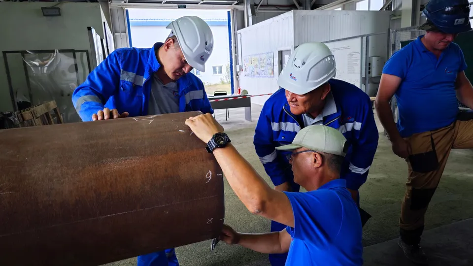Engineers in hard hats and blue uniforms examining a pipeline indoors.