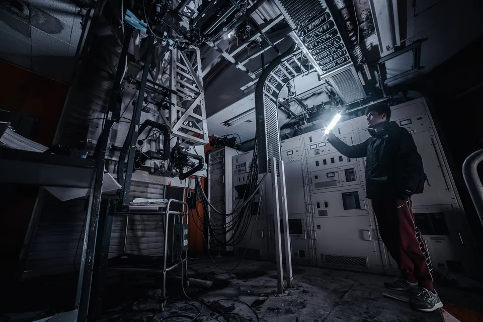 A technician using a flashlight to examine equipment in a dimly lit industrial control room.