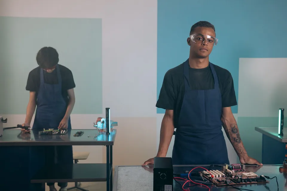 Two men working on electronics in a workshop, wearing aprons and safety goggles.